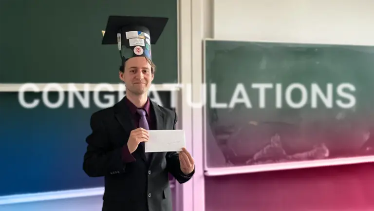 Anton Ullrich stands in front of a blackboard and smiling; the text “Congratulations” in the background is visible