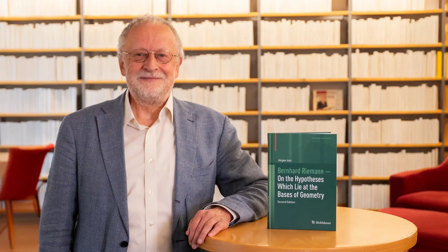 Jürgen Jost stands next to a table with his book: “Bernhard Riemann — On the Hypotheses Which Lie at the Bases of Geometry.”